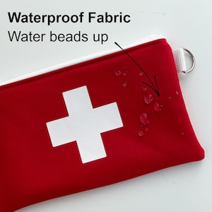 Op de afbeelding: Rode ritszak met een wit Zwitsers kruisontwerp. Water parelt op de stof, wat de waterdichte kwaliteit aantoont. De tekst "Waterproof Fabric" en "Water beads up" is zichtbaar boven de tas.