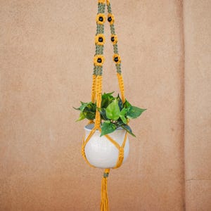 May include: A macrame plant hanger crafted from mustard yellow and olive green cord, featuring sunflower-shaped accents with black centers. The hanger supports a white ceramic pot containing a green leafy plant, finished with a long yellow tassel.