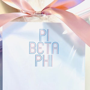 May include: A white notepad with the text "PI BETA PHI" in blue and pink, and the name "Annie Calvert" in blue script. The notepad is wrapped in clear cellophane and tied with a pink satin ribbon.