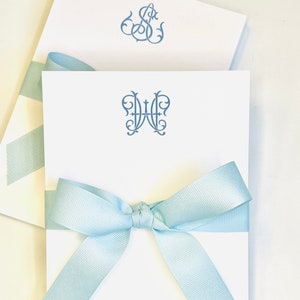 May include: White notepads with blue monogrammed initials are tied with a light blue ribbon. The top notepad features a large, ornate initial design. Another notepad is visible in the background.