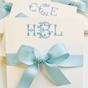 May include: A stack of square white note cards featuring a light blue monogram with the letters HBL in a decorative serif font. Each card is wrapped with a light blue grosgrain ribbon tied into a bow at the center of the front panel.