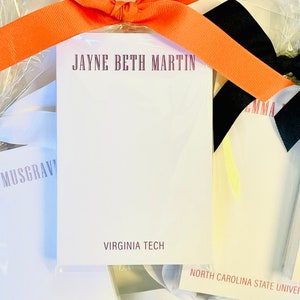 May include: A white notepad with the name "JAYNE BETH MARTIN" and "VIRGINIA TECH" printed in burgundy. The notepad is wrapped in clear cellophane and tied with a bright orange ribbon.
