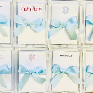 May include: Arrangement of personalized cards in clear acrylic boxes, each with a light blue ribbon bow. Names like "Christina" and "Caroline" are printed on the cards in various fonts and colors. The cards are ideal for wedding favors or personalized gifts.