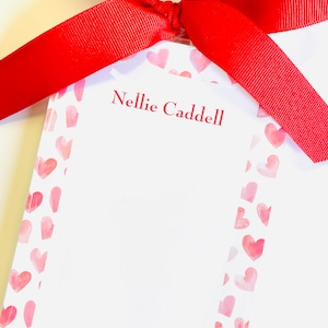 May include: A rectangular notepad featuring a white center and a border of small, light pink watercolor hearts. The name Nellie Caddell is printed in red text at the top. A bright red grosgrain ribbon is tied in a bow at the top of the notepad.