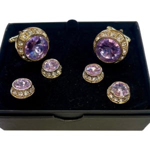 May include: A set of cufflinks and studs with a purple rhinestone center and a gold metal frame. The set is displayed in a black velvet box.