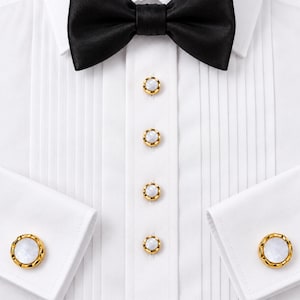 May include: A crisp white pleated tuxedo shirt featuring a black satin bow tie, four gold-rimmed buttons with white circular centers down the front placket, and matching gold-rimmed cufflinks on the cuffs of the long sleeves.