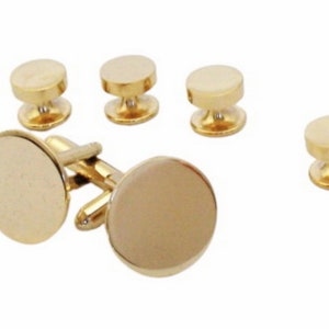 May include: A set of gold-colored cufflinks and shirt studs. The cufflinks have a circular face and a hinged backing. The shirt studs have a rounded top and a post. The set is displayed on a white background.