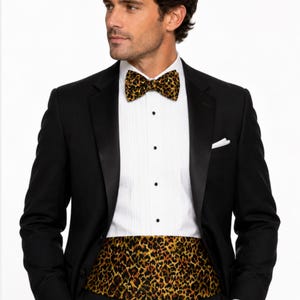 May include: A black tuxedo with a white dress shirt, black bow tie, and cummerbund featuring a leopard print pattern. The bow tie and cummerbund have a gold, brown, and black spotted design. The tuxedo has a black satin lapel.
