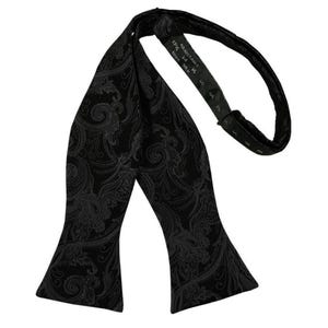 May include: A black silk bow tie featuring a tonal dark grey paisley pattern. The adjustable neck strap includes a tag printed with the text 100% SILK, ADJUSTABLE, and 14. The fabric has a subtle sheen, and the design consists of intricate swirls.