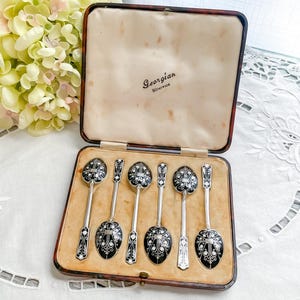 Könnte beinhalten: Ein Set aus sechs antiken Silberlöffeln mit schwarzen Emaille-Details, präsentiert in einer Vintage-Box. Die Löffel haben verzierte Designs auf den Griffen und Schalen. Die Box ist braun mit cremefarbenem Innenraum.