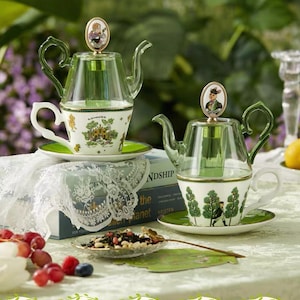 Puede incluir: Dos teteras de vidrio con detalles verdes se asientan sobre tazas y platillos a juego. Las tazas presentan un diseño caprichoso con ilustraciones verdes y blancas. Un pequeño plato de hojas de té y fruta está en primer plano.