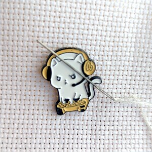 May include: A white cat wearing headphones and holding a video game controller. The cat is a cartoon-style enamel pin.