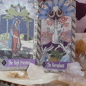 Puede incluir: Dos cartas de tarot, La Sacerdotisa y El Hierofante, se muestran sobre una superficie blanca con cristales. La carta de La Sacerdotisa presenta a una mujer con una túnica azul y una falda roja de pie en un bosque. La carta del Hierofante presenta a un hombre con una túnica blanca de pie en un bosque con un árbol detrás de él.