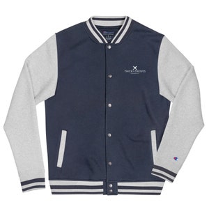 May include: A navy blue and grey varsity jacket with a white embroidered logo that says "THICK & THIEVES UNIVERSITY" and a white "X" above the text. The jacket has a zipper closure and a ribbed collar, cuffs, and hem.