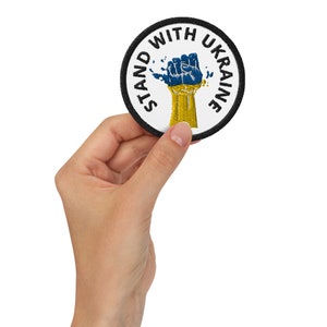 May include: A round, white patch with black text that reads "Stand With Ukraine". A blue and yellow fist is in the centre of the patch.