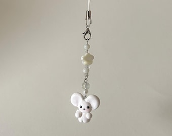 May include: A white mouse charm with a silver-tone clasp and a string. The charm features a small, cartoon-like mouse figure with large ears and a cute face. The charm is adorned with white beads and a star-shaped bead.