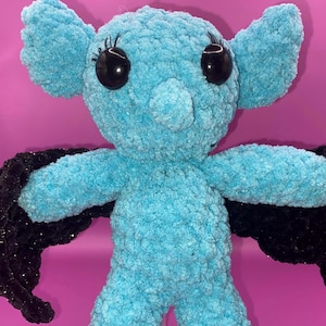 Puede incluir: Una muñeca de murciélago de peluche azul hecha a mano. El murciélago tiene grandes ojos negros con pestañas, una nariz pequeña y orejas grandes. Tiene alas negras y está hecho de un material suave y texturizado. La muñeca está sobre un fondo rosa.