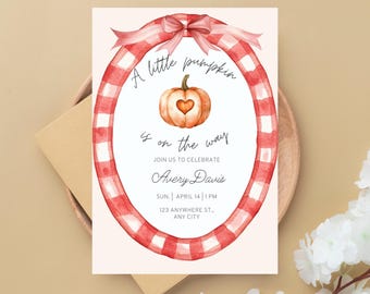 May include: A baby shower invitation with a watercolor design. The invitation features a pumpkin with a heart-shaped cutout, surrounded by a red and white gingham oval frame. The text reads "A little pumpkin is on the way."