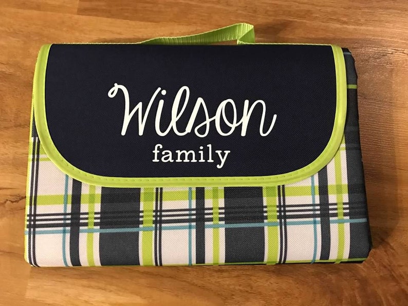 Puede incluir: Una manta de picnic de cuadros azules y verdes con una solapa azul marino que tiene el texto "Wilson family" en letras blancas cursivas. La manta tiene un asa de transporte.