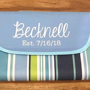 Puede incluir: Una manta de picnic a rayas azules y blancas con una solapa azul claro que tiene el texto "Becknell Est. 7/16/18" en blanco.