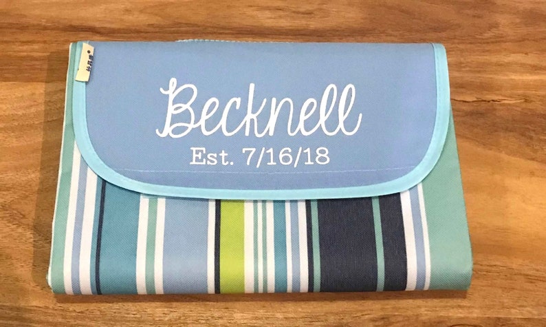 Puede incluir: Una manta de picnic a rayas azules y blancas con una solapa azul claro que tiene el texto "Becknell Est. 7/16/18" en blanco.
