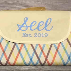 Puede incluir: Una manta de picnic de color crema con un patr&oacute;n de cuadros marrones, azules, naranjas y amarillos. La manta tiene una solapa con un fondo de color crema y una inscripci&oacute;n azul que dice "Seel Est. 2019".