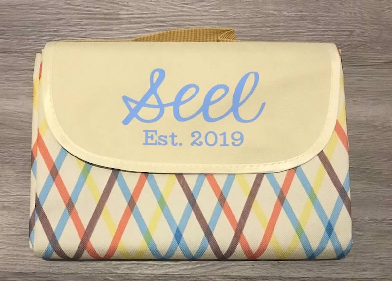 Puede incluir: Una manta de picnic de color crema con un patr&oacute;n de cuadros marrones, azules, naranjas y amarillos. La manta tiene una solapa con un fondo de color crema y una inscripci&oacute;n azul que dice "Seel Est. 2019".