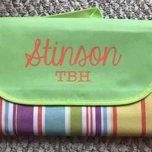 Puede incluir: Una manta de picnic a rayas verdes y blancas con una solapa verde. La solapa tiene el nombre "Stinson" y las iniciales "TBH" bordadas en naranja.