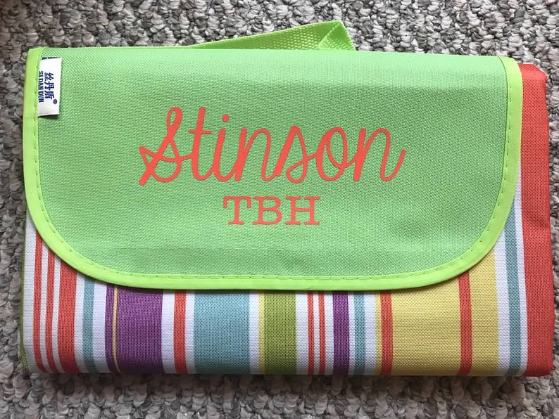 Puede incluir: Una manta de picnic a rayas verdes y blancas con una solapa verde. La solapa tiene el nombre "Stinson" y las iniciales "TBH" bordadas en naranja.