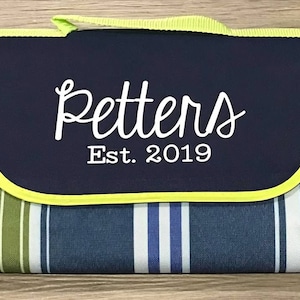 Puede incluir: Una manta de picnic a rayas azules y verdes con una solapa azul marino. La solapa tiene un borde amarillo y texto blanco que dice "Petters Est. 2019".