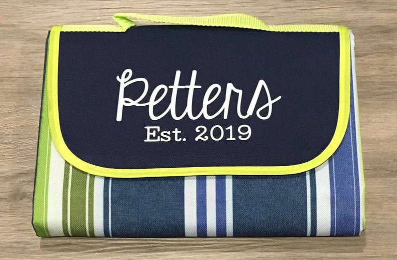 Puede incluir: Una manta de picnic a rayas azules y verdes con una solapa azul marino. La solapa tiene un borde amarillo y texto blanco que dice "Petters Est. 2019".
