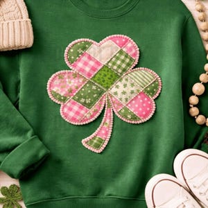 May include: Green sweatshirt featuring a large, patchwork shamrock design in pink and green. The shamrock is outlined with a pink beaded trim. A beige knit hat and white sneakers are visible in the image.