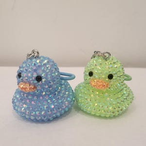 May include: Two bejeweled duck keychains. One is light blue, the other is light green. Both have black eyes and orange beaks. Each duck has a silver keychain ring and a matching colored loop.