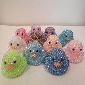 May include: A collection of bedazzled chick figurines in various colors, including pink, blue, green, and purple. Each chick has a small orange beak and black eyes. The figurines are clustered together, creating a playful and decorative display.