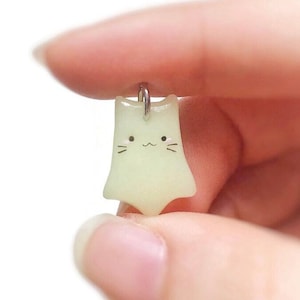 以下が含まれることがあります： 銀色のループが付いた、小さく光る猫の形をしたペンダント。ペンダントには、黒いひげと笑顔の口のかわいい顔があります。猫は柔らかい淡い緑色です。