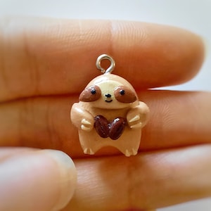 May include: A small, brown clay sloth charm with a heart in its hands. The charm has a silver loop for hanging.