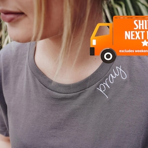 A person wearing a gray t-shirt with the word 'pray' written on it, standing in front of a grassy background with an orange shipping truck in the background.