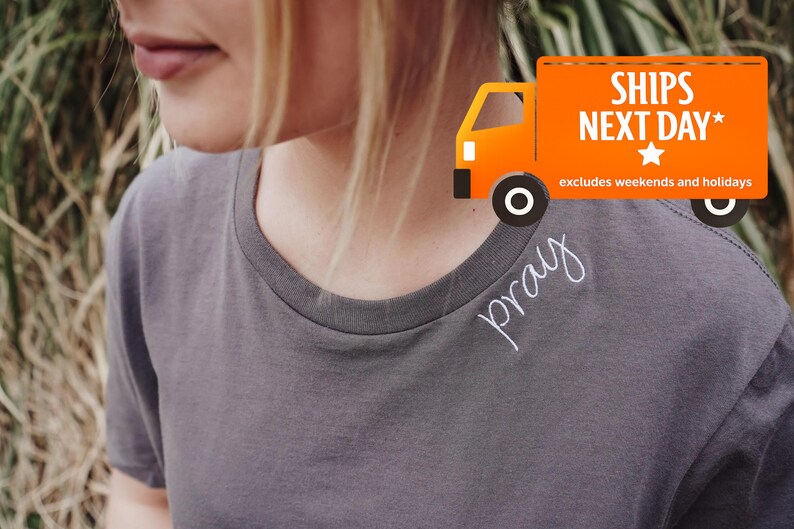 A person wearing a gray t-shirt with the word 'pray' written on it, standing in front of a grassy background with an orange shipping truck in the background.
