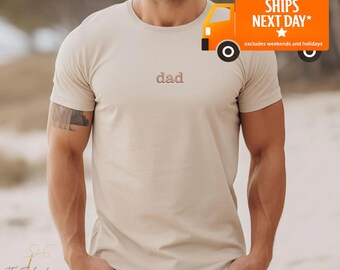 Puede incluir: Camiseta beige de cuello redondo con la palabra "dad" bordada en hilo marrón en el pecho. La camiseta tiene mangas cortas y está hecha de un material suave. Un pequeño gráfico de camión naranja con el texto "SHIPS NEXT DAY" es visible.