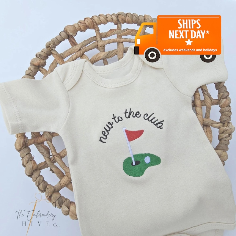 A white baby onesie with a golf hole embroidered on it, placed on a woven basket. There is a shipping truck sticker on the basket, indicating that the item is being shipped next day.