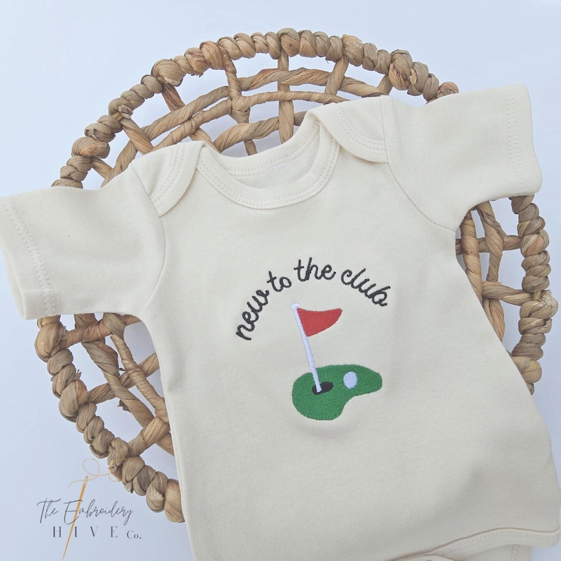 A white baby onesie with the text "new to the club" and a golf hole embroidered on it, placed on a woven basket.