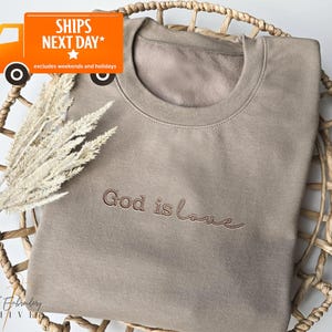 May include: A tan-colored sweatshirt with the embroidered phrase "God is love" in a cursive font. The sweatshirt is displayed in a woven basket with dried decorative plants. The text "SHIPS NEXT DAY" is in the upper left corner.