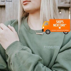 May include: Sage green sweatshirt with the word "custom" embroidered on the front. A silver bar necklace is worn. An orange truck graphic with the text "SHIPS NEXT DAY" is in the background.