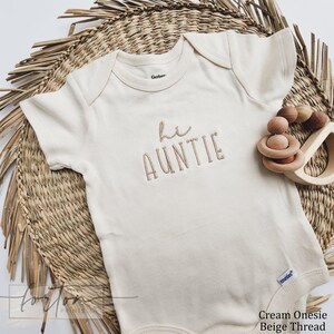 A cream-colored onesie with the text "Hi Auntie" embroidered on it, placed on a woven mat and accompanied by wooden toys.
