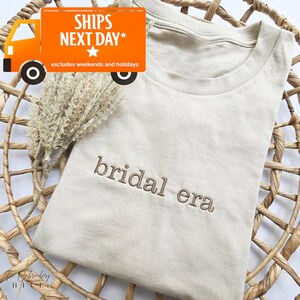 May include: A beige t-shirt with the embroidered text "bridal era" in a brown thread. The shirt is folded and placed on a woven basket. The image also includes a sprig of dried flowers and a graphic that says "SHIPS NEXT DAY".