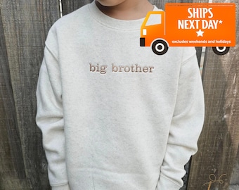 May include: A cream-colored crewneck sweatshirt featuring the text big brother embroidered in a brown thread across the chest. The garment has long sleeves with ribbed cuffs and a ribbed neckline, constructed from a speckled fabric material.