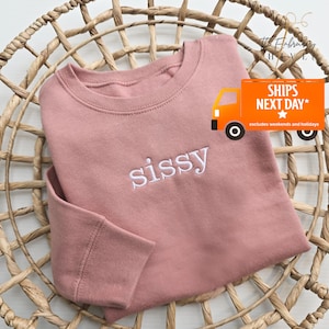 May include: A dusty rose-colored sweatshirt with the word "sissy" embroidered in white. The sweatshirt is folded and placed in a woven basket. The text "SHIPS NEXT DAY" is visible on the right side of the image.