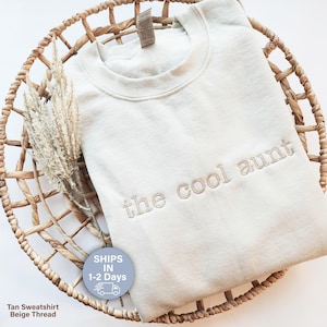 May include: A tan-colored sweatshirt with the embroidered text "the cool aunt" in a matching beige thread. The sweatshirt is displayed in a woven basket with dried decorative grass. The text "Tan Sweatshirt Beige Thread" is at the bottom.
