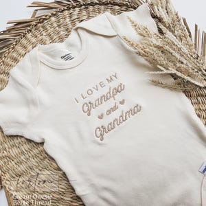 A white baby onesie with the text "I LOVE MY GRANDPA AND GRANDMA" embroidered on it, placed on a woven basket.
