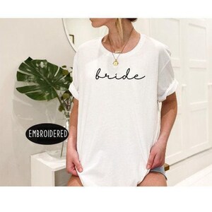 A woman wearing a white t-shirt with the word "bride" printed on it, standing in front of a mirror and a plant.